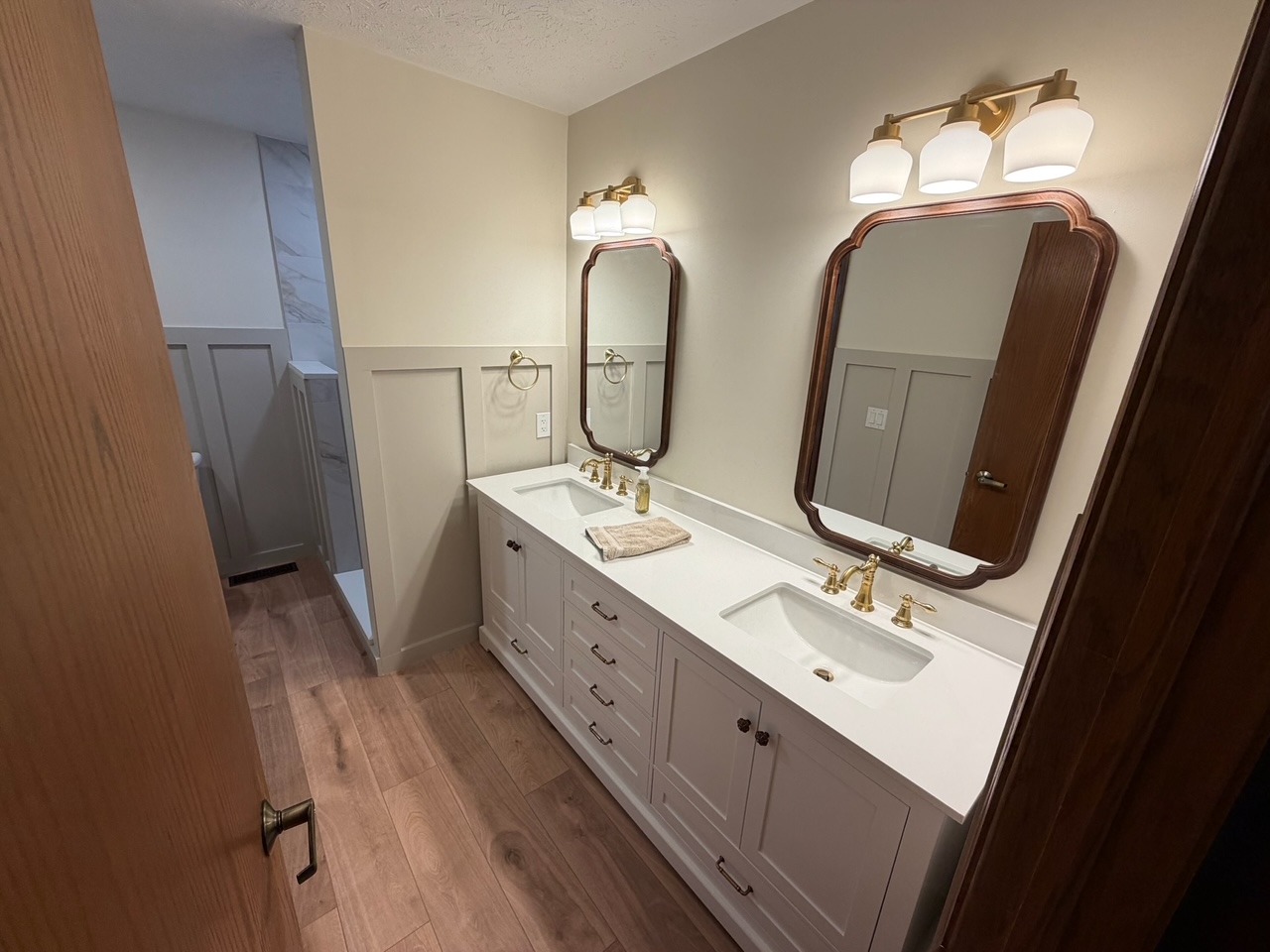 Completed double vanity bathroom remodel with brass fixtures, framed mirrors and wood-look flooring